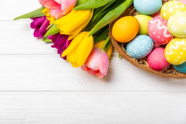 Renkli yumurtalarla dolu Paskalya sepeti ve ahşap bir masanın üzerinde bir buket lale. Paskalya kutlaması. El yapımı renkli Paskalya yumurtaları. Mesaj için yer. Boşluğu kopyala