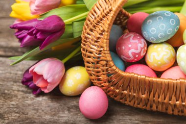 Renkli yumurtalarla dolu Paskalya sepeti ve ahşap bir masanın üzerinde bir buket lale. Paskalya kutlaması. El yapımı renkli Paskalya yumurtaları. Mesaj için yer. Boşluğu kopyala