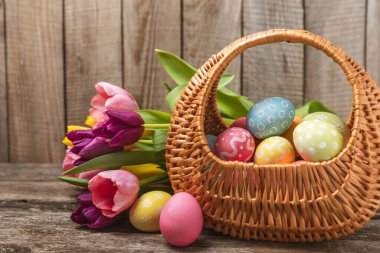 Renkli yumurtalarla dolu Paskalya sepeti ve ahşap bir masanın üzerinde bir buket lale. Paskalya kutlaması. El yapımı renkli Paskalya yumurtaları. Mesaj için yer. Boşluğu kopyala