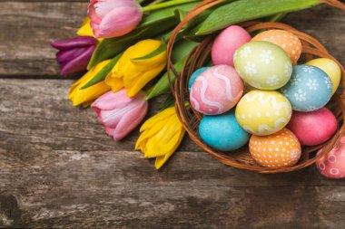 Renkli yumurtalarla dolu Paskalya sepeti ve ahşap bir masanın üzerinde bir buket lale. Paskalya kutlaması. El yapımı renkli Paskalya yumurtaları. Mesaj için yer. Boşluğu kopyala