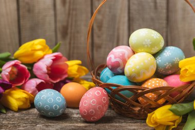 Renkli yumurtalarla dolu Paskalya sepeti ve ahşap bir masanın üzerinde bir buket lale. Paskalya kutlaması. El yapımı renkli Paskalya yumurtaları. Mesaj için yer. Boşluğu kopyala