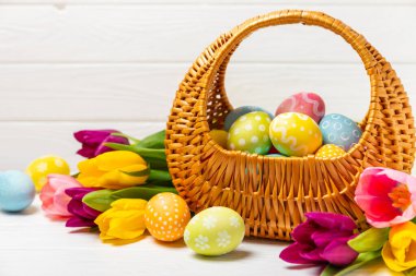Renkli yumurtalarla dolu Paskalya sepeti ve ahşap bir masanın üzerinde bir buket lale. Paskalya kutlaması. El yapımı renkli Paskalya yumurtaları. Mesaj için yer. Boşluğu kopyala