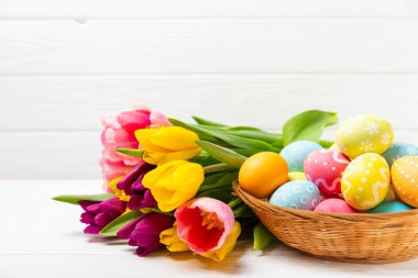 Renkli yumurtalarla dolu Paskalya sepeti ve ahşap bir masanın üzerinde bir buket lale. Paskalya kutlaması. El yapımı renkli Paskalya yumurtaları. Mesaj için yer. Boşluğu kopyala