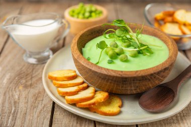 Ahşap bir masada yeşil bezelye çorbası. Taze bezelye çorbası kasesi. Yeşil bezelye ve kızarmış ekmekle dolu bir kasede lezzetli, sağlıklı krema çorbası. Sağlıklı vejetaryen püresi. Uzayı kopyala Metin için boşluk.