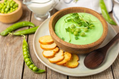 Ahşap bir masada yeşil bezelye çorbası. Taze bezelye çorbası kasesi. Yeşil bezelye ve kızarmış ekmekle dolu bir kasede lezzetli, sağlıklı krema çorbası. Sağlıklı vejetaryen püresi. Uzayı kopyala Metin için boşluk.