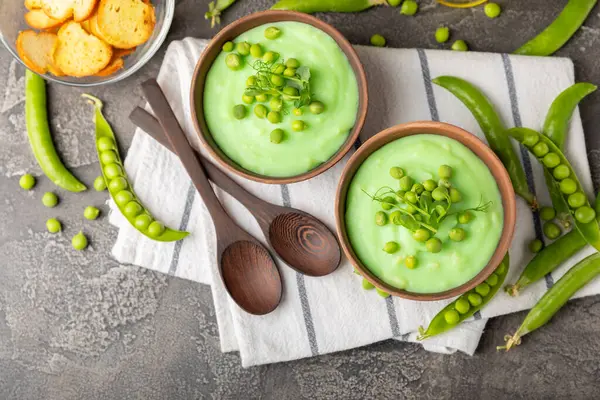 Ahşap bir masada yeşil bezelye çorbası. Taze bezelye çorbası kasesi. Yeşil bezelye ve kızarmış ekmekle dolu bir kasede lezzetli, sağlıklı krema çorbası. Sağlıklı vejetaryen püresi. Uzayı kopyala Metin için boşluk.