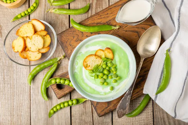 Ahşap bir masada yeşil bezelye çorbası. Taze bezelye çorbası kasesi. Yeşil bezelye ve kızarmış ekmekle dolu bir kasede lezzetli, sağlıklı krema çorbası. Sağlıklı vejetaryen püresi. Uzayı kopyala Metin için boşluk.