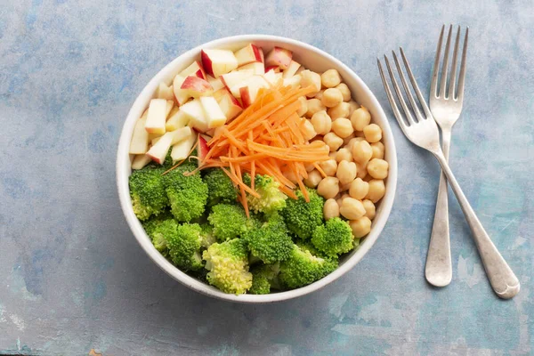 Broccoli salad add apple chickpeas in white bowl isolated on blue wood background close up, top view, healthy food concept.