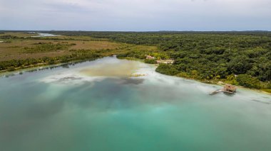 Bacalar, Mexico, Quintana Roo, Beautiful blue lake with a small island in the middle. The island is surrounded by trees and there are some houses on it. The sky is cloudy and the water is calm
