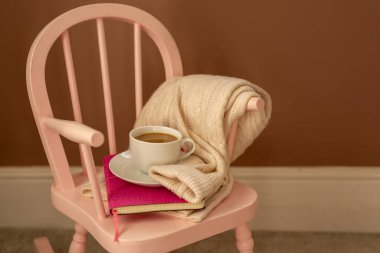 Childrens room, leisure, coziness, hygge. Pink childrens rocking chair against the wall with delicious cocoa in a white mug on a saucer, a knitted sweater and a book clipart