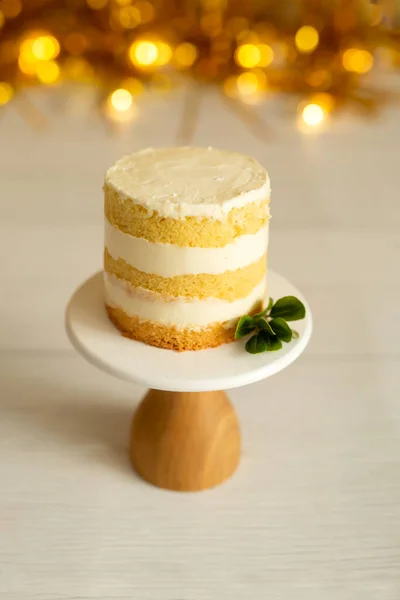 Celebration. A small cake, bare cakes of dough and white cream, on a brown wooden stand on a blurred background of gold decor. Selective focus