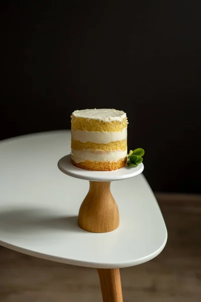 Holiday, celebration. A small cake, bare cakes of dough and white cream, with eucalyptus on a brown wooden stand on a black background. Selective focus