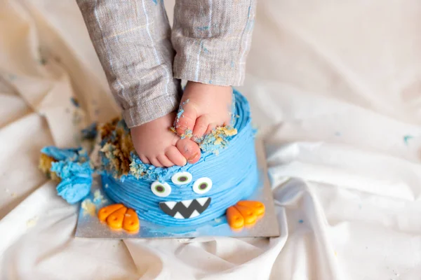 First birthday cake smash fun. Bare feet of a boy in gray pants on a blue cake in the form of a freaks face