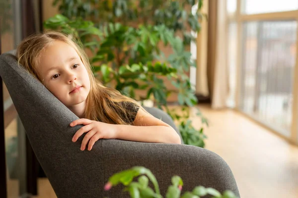 Rest, child development, plants at home. A cute girl with long blond hair in a gray chair on the terrace against the background of green plants.