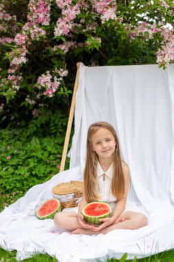 Piknik, yaz, atıştırmalık. Beyaz elbiseli şirin bir kız yeşil çimenli ve çiçek açan bir ağaçlı bir bahçede beyaz bir battaniyenin üzerinde oturuyor. Piknik sepeti ve karpuz.