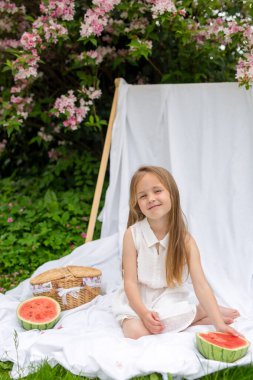 Piknik, yaz, atıştırmalık. Beyaz elbiseli şirin bir kız yeşil çimenli ve çiçek açan bir ağaçlı bir bahçede beyaz bir battaniyenin üzerinde oturuyor. Piknik sepeti ve karpuz.