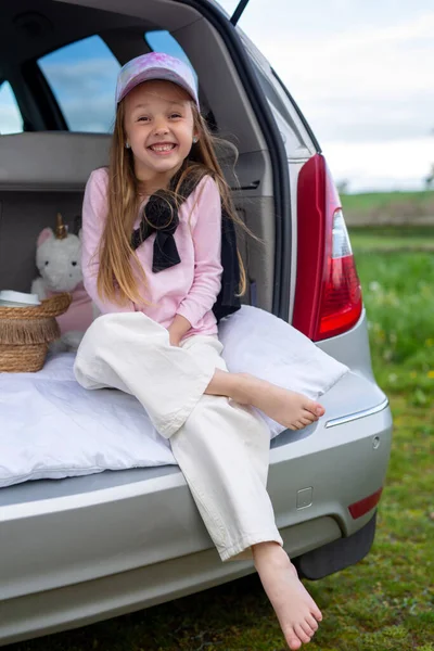 Traveling by car, rest, childhood. A cute girl in a cap in stylish clothes sits in the trunk of a gray car.