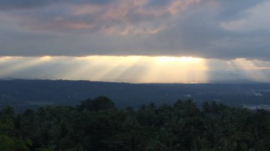 Yoğun karanlık bulutları delip geçen, tropikal ormanları ve uzak dağları aşan güneş ışınlarıyla dramatik bir manzara. Umut, barış, doğa ve maneviyat teması için ideal..