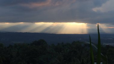 Yoğun karanlık bulutları delip geçen, tropikal ormanları ve uzak dağları aşan güneş ışınlarıyla dramatik bir manzara. Umut, barış, doğa ve maneviyat teması için ideal..