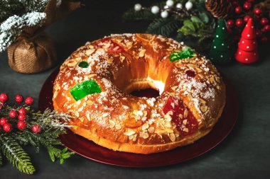 Kremalı Roscon de Reyes ve gri bir masadaki kırmızı tabakta noel süsleri. Krallar Günü konsepti İspanyolca üç papaz pastası. Noel için tipik İspanyol tatlısı.