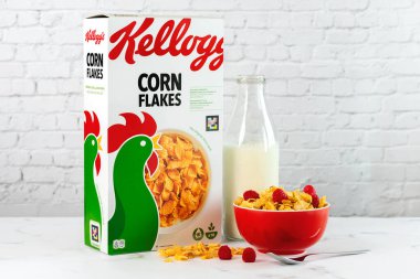 Mısır gevreği ve ahududu dolu kırmızı bir kase ve mermer bir masada süt şişesi ile Kellogg 'un kutusu.