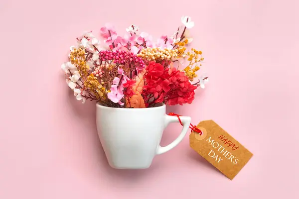 Happy Mother\'s Day. White cup of coffee with various flowers and brown sale tag with the text Happy Mothers day over pink background