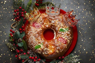 Kırmızı bir tabakta Roscon de Reyes 'in üst görüntüsü kırmızı böğürtlen dalları ve Noel ışıklarının arka planında bilge adamların taçlarıyla. Krallar Günü konsepti İspanyolca üç kralın pastası