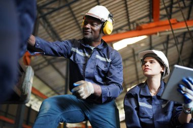 Young successful technician or repairman of industrial machines in safety helmet and workwear standing in large garage or workshop