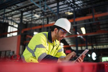 Young successful technician or repairman of industrial machines in safety helmet and workwear standing in large garage or workshop