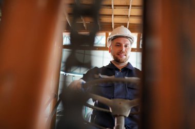 Young successful technician or repairman of industrial machines in safety helmet and workwear standing in large garage or workshop