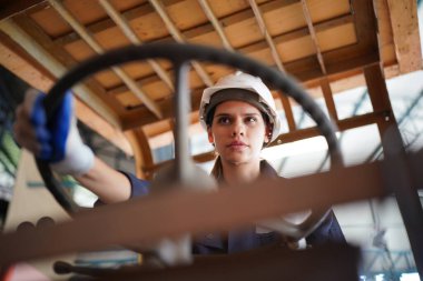 Young successful technician or repairman of industrial machines in safety helmet and workwear standing in large garage or workshop