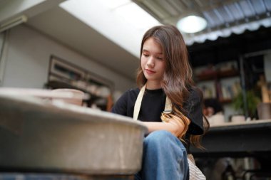 Konsantre olmuş, önlüklü, çömlek direksiyonunda oturan ve ıslak kil kabı şekillendirirken el işi aletleri kullanan güzel bir zanaatkar.