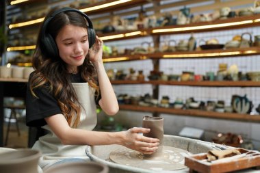 Konsantre olmuş, önlüklü, çömlek direksiyonunda oturan ve ıslak kil kabı şekillendirirken el işi aletleri kullanan güzel bir zanaatkar.