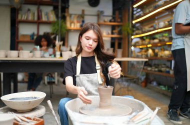 Konsantre olmuş, önlüklü, çömlek direksiyonunda oturan ve ıslak kil kabı şekillendirirken el işi aletleri kullanan güzel bir zanaatkar.