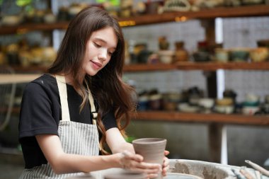 Konsantre olmuş, önlüklü, çömlek direksiyonunda oturan ve ıslak kil kabı şekillendirirken el işi aletleri kullanan güzel bir zanaatkar.