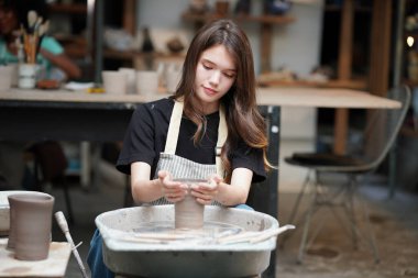 Konsantre olmuş, önlüklü, çömlek direksiyonunda oturan ve ıslak kil kabı şekillendirirken el işi aletleri kullanan güzel bir zanaatkar.