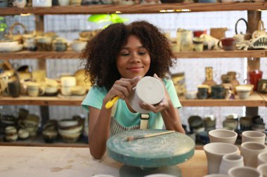 Konsantre olmuş, önlüklü, çömlek direksiyonunda oturan ve ıslak kil kabı şekillendirirken el işi aletleri kullanan güzel bir zanaatkar.