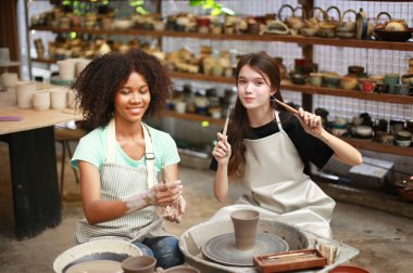Konsantre olmuş, önlüklü, çömlek direksiyonunda oturan ve ıslak kil kabı şekillendirirken el işi aletleri kullanan güzel bir zanaatkar.