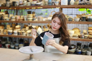 Konsantre olmuş, önlüklü, çömlek direksiyonunda oturan ve ıslak kil kabı şekillendirirken el işi aletleri kullanan güzel bir zanaatkar.