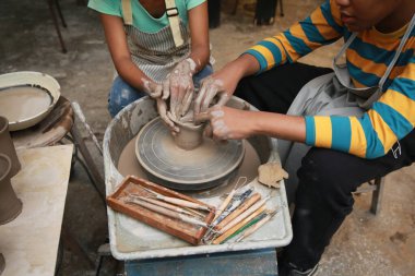 Konsantre olmuş, önlüklü, çömlek direksiyonunda oturan ve ıslak kil kabı şekillendirirken el işi aletleri kullanan güzel bir zanaatkar.