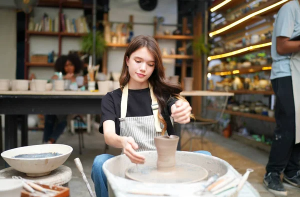 Konsantre olmuş, önlüklü, çömlek direksiyonunda oturan ve ıslak kil kabı şekillendirirken el işi aletleri kullanan güzel bir zanaatkar.
