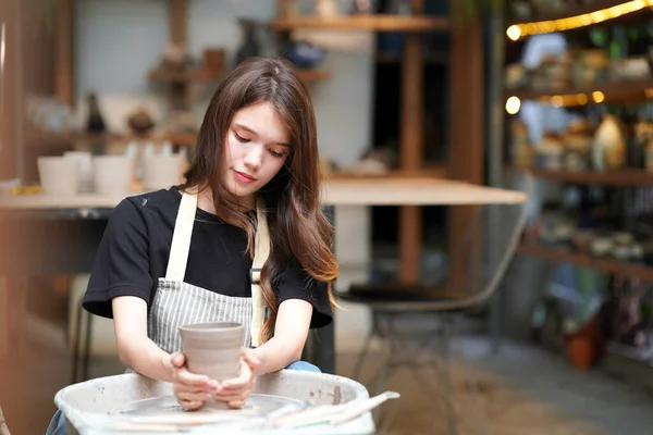 Close-up of concentrated beautiful craftswoman in apron sitting at pottery wheel and using craft tool while shaping wet clay vessel