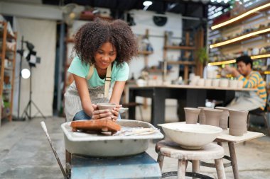Konsantre olmuş, önlüklü, çömlek direksiyonunda oturan ve ıslak kil kabı şekillendirirken el işi aletleri kullanan güzel bir zanaatkar.