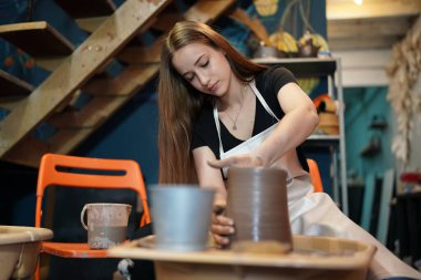 Konsantre olmuş, önlüklü, çömlek direksiyonunda oturan ve ıslak kil kabı şekillendirirken el işi aletleri kullanan güzel bir zanaatkar.
