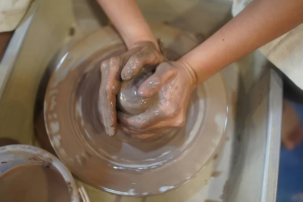 Close up view of craftswoman in apron sitting at pottery wheel and using craft tool while shaping wet clay vessel