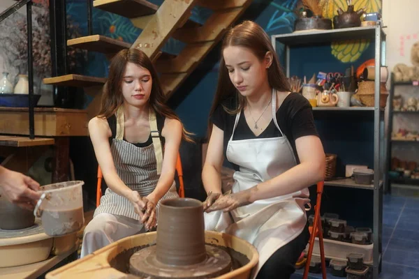 Close-up of concentrated beautiful craftswoman in apron sitting at pottery wheel and using craft tool while shaping wet clay vessel