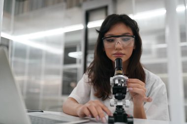 Laboratuvarda mikroskopla çalışan üniformalı genç kadın doktor analiz yapıyor.