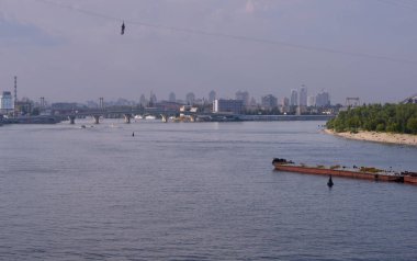 Dnipro nehrinde yüzen büyük bir mavna, Truhanov adası kumlu sahilinde insanlar arka planda güneşleniyor..