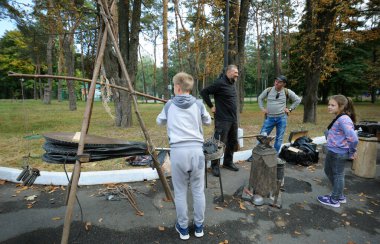 Kalpazanlık öğretiyorum. Çocuk körük kullanarak demir ocağına hava pompalıyor, küçük kız ve izleyen bir adam. 28 Eylül 2019. Kiev, Ukrayna