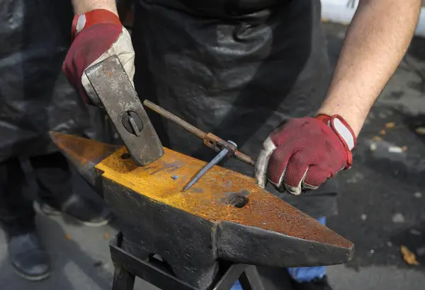 Blacksmith hands holding forceps and a hammer forging a metal billet nail on an anvil.